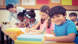 Grundschulkinder sitzen an ihren Tischen und lernen. Die Lehrerin steht am Ende der Reihe und beugt sich zur Hilfe über eine Schülerin.