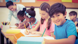 Grundschulkinder sitzen an ihren Tischen und lernen. Die Lehrerin steht am Ende der Reihe und beugt sich zur Hilfe über eine Schülerin.