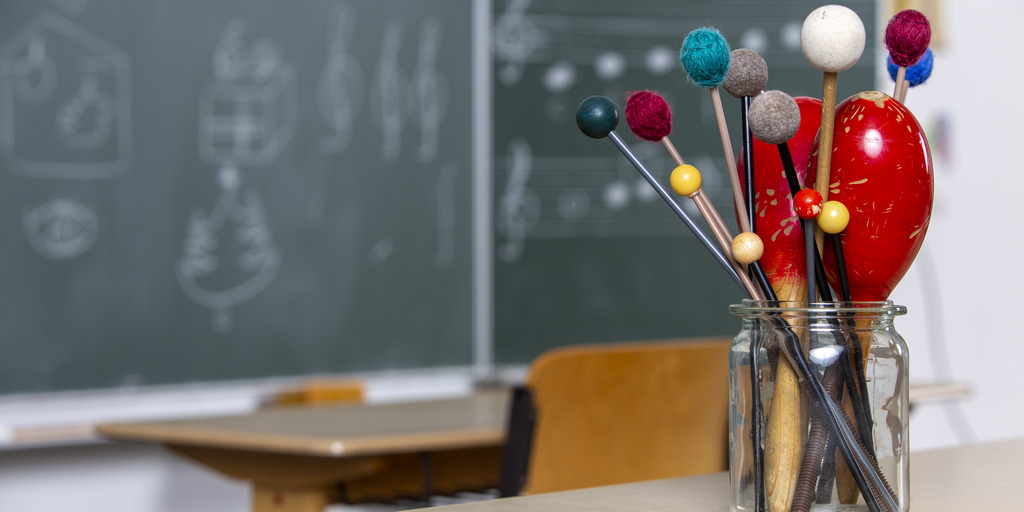 Fotoaufnahme eines Musik-Klassenzimmers. Im Vordergrund steht ein durchsichtiges Glasgefäß mit Schlägeln und Maracas. Im Hintergrund ist eine Tafel zu sehen.