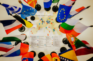 Fähnchen verschiedener europäischer Staaten stehen kreisförmig um eine Europakarte, auf der die Standorte der Bürgerdialoge im Rahmen des Projekts "From local to European" markiert sind.