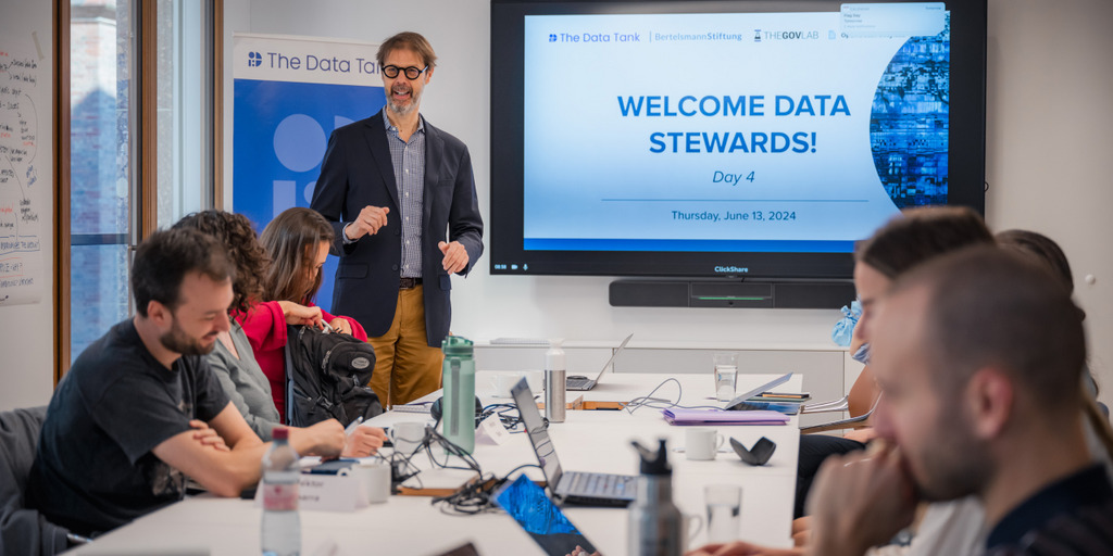 In einem Raum steht vorne neben einem großen Monitor Stefaan Verhulst. An den Tischen sitzen Teilnehmerinnen und Teilnehmer des Kurses. Auf dem Monitor steht: Welcome Data Stewards.