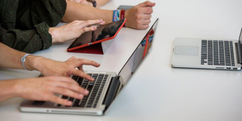 Drei Schüler, von denen nur die Hände und Arme zu sehen sind, sitzen an einem Tisch und arbeiten mit einem Laptop und einem Tablet-PC.