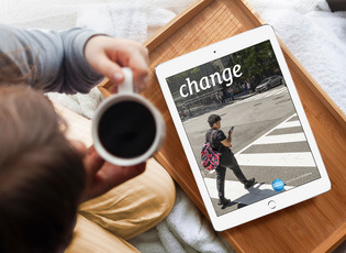 Eine Frau sitzt mit der Kaffeetasse in der Hand vor einem Tablett, auf dem ein Tablet-PC liegt, auf dem das Titelbild der aktuellen change-Ausgabe zu sehen ist. Das Titelbild zeigt einen jungen Mann, der in Washington, DC einen Zebrastreifen überquert und dabei auf sein Handy guckt.