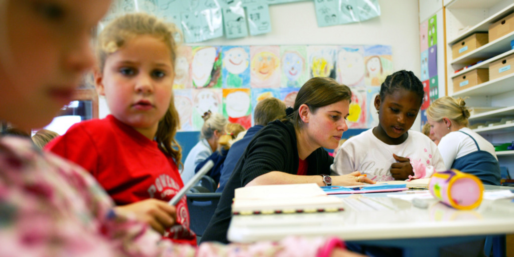 Eine Lehrerin liest im Klassenzimmer mit einer Schülerin in einem Buch. Rings umher sitzen weitere Schüler und Lehrerinnen.