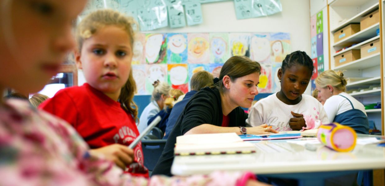 (© Veit Mette, Bielefeld) Eine Lehrerin liest im Klassenzimmer mit einer Schülerin in einem Buch. Rings umher sitzen weitere Schüler und Lehrerinnen.