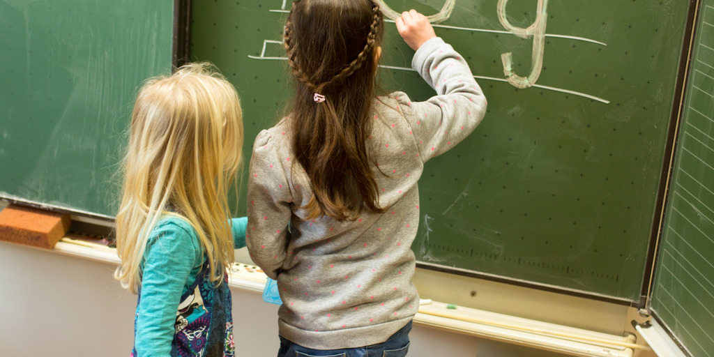 Zwei Mädchen stehen vor einer Schultafel in einem Klassenzimmer. Eins der Mädchen schreibt mit Kreide an die Tafel.