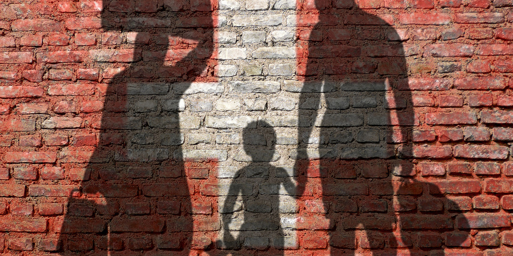 Backsteinmauer in den Farben der schweizerischen Flagge. Darauf zu sehen die Schatten einer Frau, eines Jungen und eines Mannes.