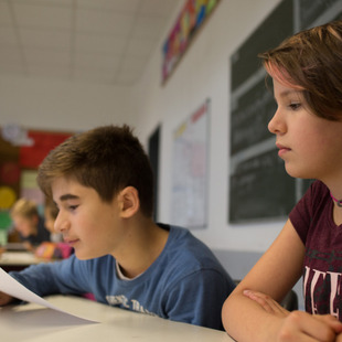Ein etwas 11jähriger Schüler liest etwas von einem Blatt ab, neben ihm sitzt eine Schülerin