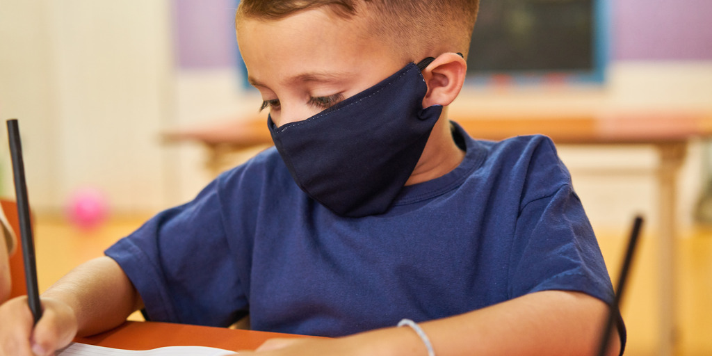 Junge mit Mundschutz sitzt in der Schule am Tisch und schreibt