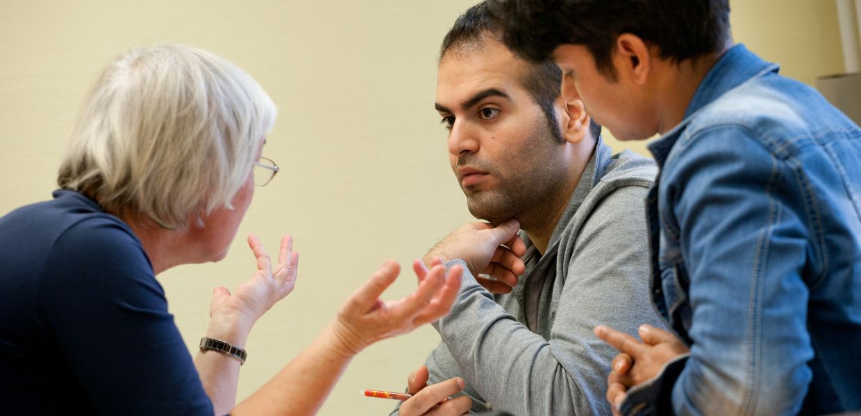 (© Thomas Kunsch) An older woman is talking with two young men during a German lesson for immigrants.