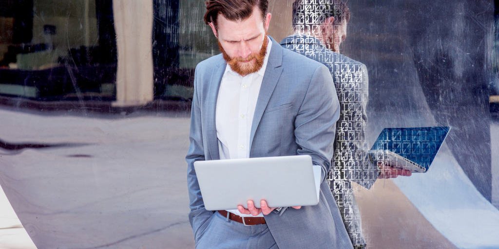 Ein junger Geschäftsmann im Anzug steht vor einer Glaswand und blickt auf einen Laptop, den er auf der linken Hand balanciert. Hinter ihm sind auf die Glaswand Nullen und Einsen projiziert, also Symbole für eine Programmiersprache.