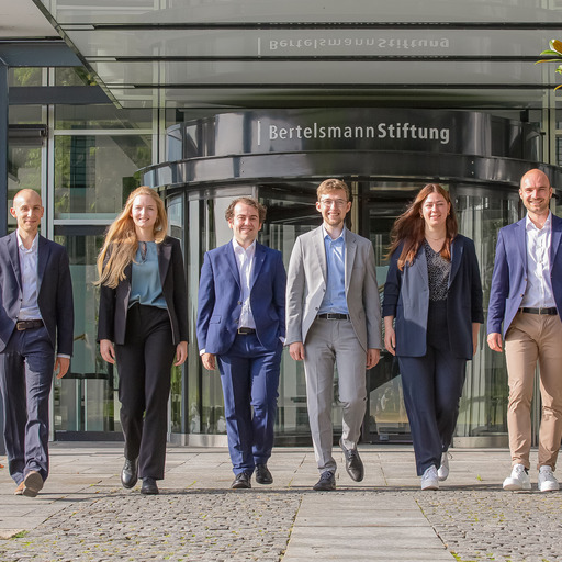 Die sechs Junior Professionals der Bertelsmann Stiftung des Jahrgangs 2023 - Jakob Weber, Anne Meisiek, Etienne Höra, Leander Berner, Lena Budach und Otto Meyer zu Schwabedissen - gehen gemeinsam den Weg vor dem Eingang zur Bertelsmann Stiftung hinunter und blicken in die Kamera.
