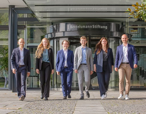 Die sechs Junior Professionals der Bertelsmann Stiftung des Jahrgangs 2023 - Jakob Weber, Anne Meisiek, Etienne Höra, Leander Berner, Lena Budach und Otto Meyer zu Schwabedissen - gehen gemeinsam den Weg vor dem Eingang zur Bertelsmann Stiftung hinunter und blicken in die Kamera.