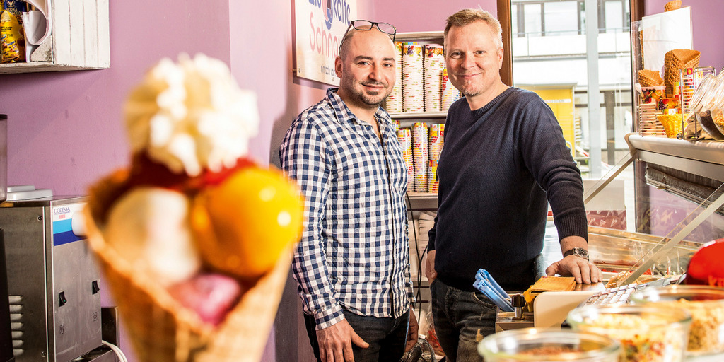 Zwei Männer stehen hinter der Theke in einem Eiscafé. Im Vordergrund links steht eine mit Kugeln gefüllte Eiswaffel.