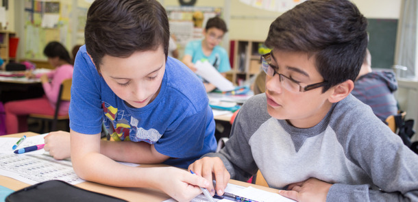 Zwei Jungs sitzen nebeneinander, ein Junge erklärt dem anderen etwas, sie schauen in ein SchulheftZwei Jungs sitzen nebeneinander, ein Junge erklärt dem anderen etwas, sie schauen in ein Schulheft