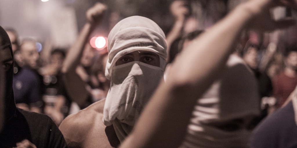 A protester at an anti-government rally in the Lebanese capital of Beirut is wearing his t-shirt as a mask in order to protect himself against tear gas. Next to and behind him, other protesters have raised their arms.