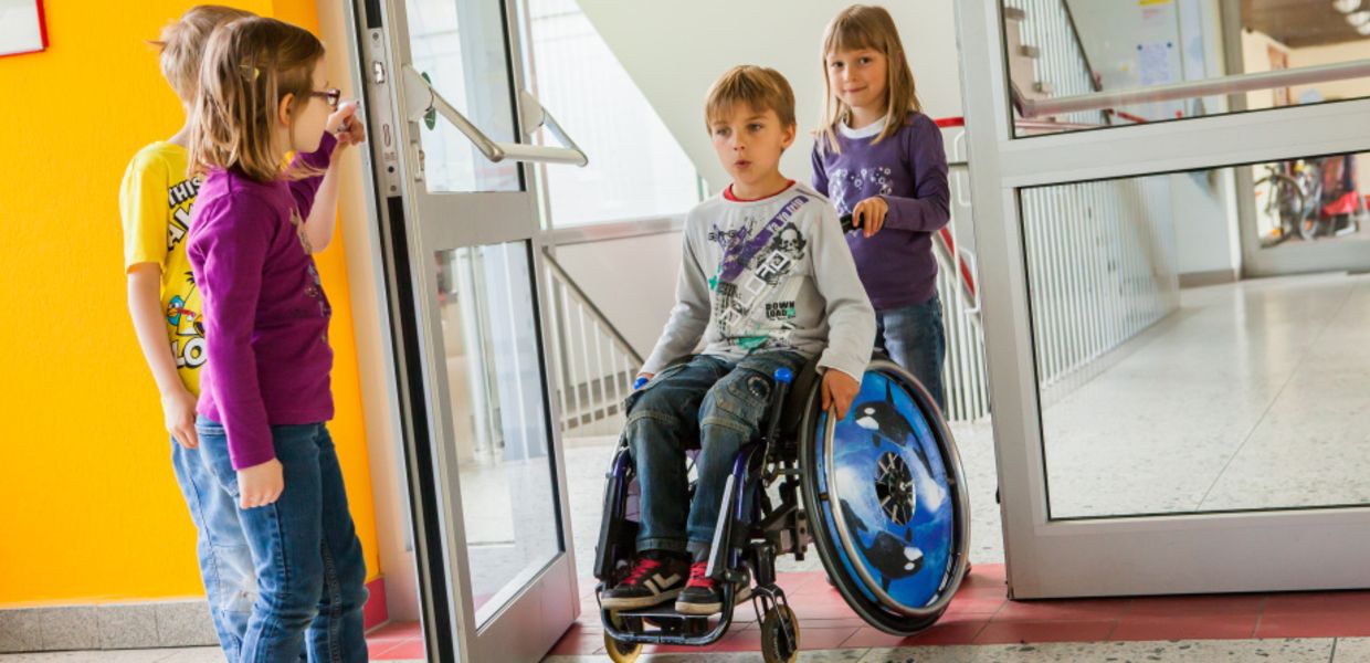 (© Jan Voth) Zwei Kinder halten in einer Grundschule die Tür auf, während ein Mädchen einen Jungen im Rollstuhl durch die Tür schiebt.