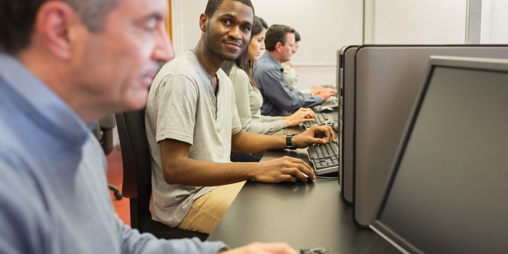 Several people are sitting in a computer class. One man looks up from the monitor and directly into the camera.