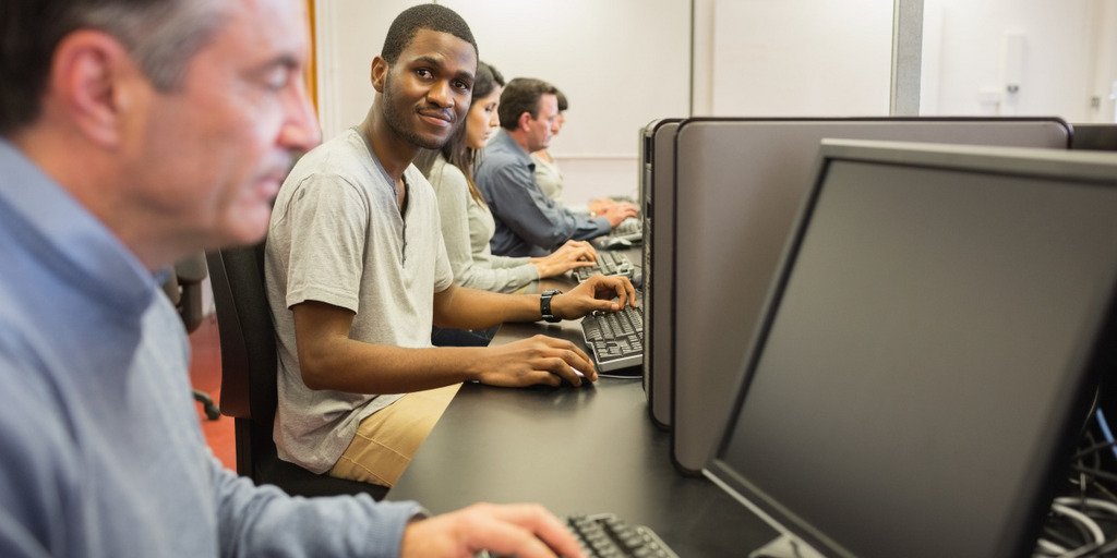 Mehrere Menschen sitzen in einem Computer-Kurs - eine Person schaut vom Bildschirm hoch in die Kamera.