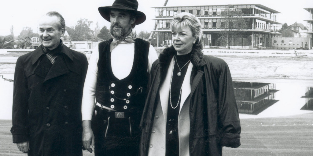 Reinhard Mohn steht mit seiner Frau Liz Mohn und dem Zimmermann Wilhelm Boeger vor dem Neubau des Stiftungsgebäudes