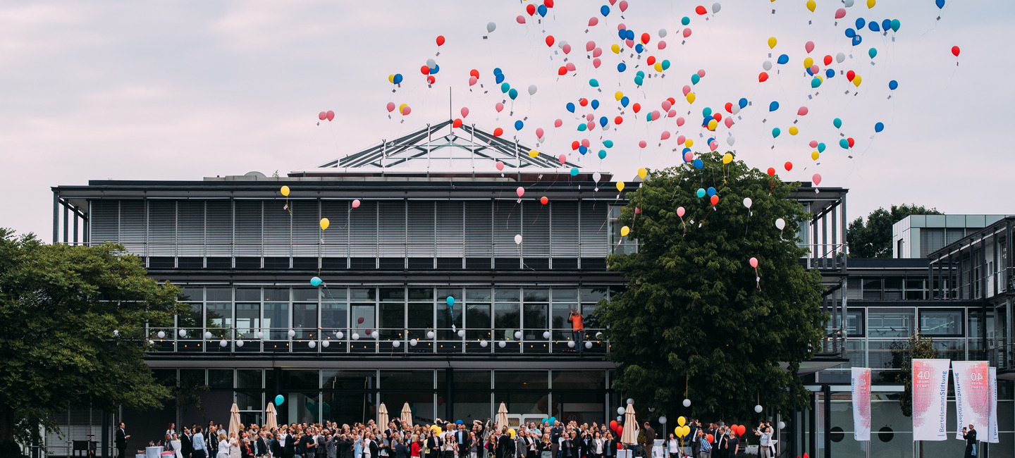 Blick über den See vor dem Gebäude auf das Gebäude der Bertelsmann Stiftung während eines Sommerfestes. Bunte Luftballons steigen von der Terrasse vor dem Gebäude auf, auf der die Mitarbeiter:innen der Stiftung versammelt sind.