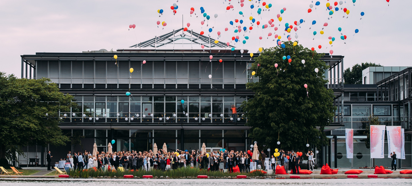 Blick über den See vor dem Gebäude auf das Gebäude der Bertelsmann Stiftung während eines Sommerfestes. Bunte Luftballons steigen von der Terrasse vor dem Gebäude auf, auf der die Mitarbeiter:innen der Stiftung versammelt sind.