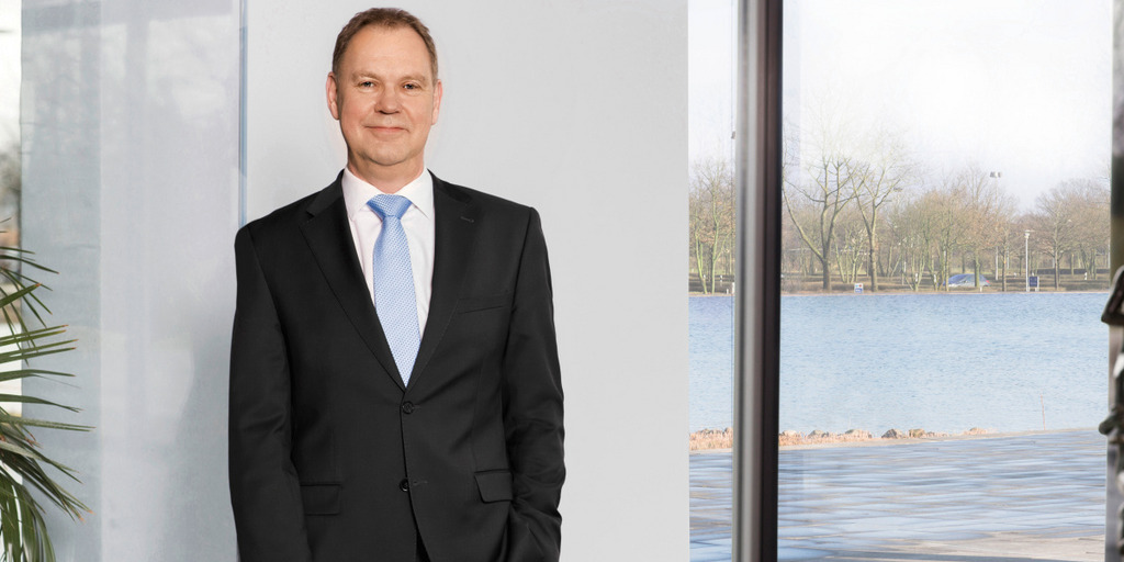 Aart De Geus, Vorstandsvorsitzender der Bertelsmann Stiftung, steht in einem Raum der Stiftung vor einer Wand und blickt in die Kamera. Im Hintergrund eine Pflanze, Fenster und ein See.