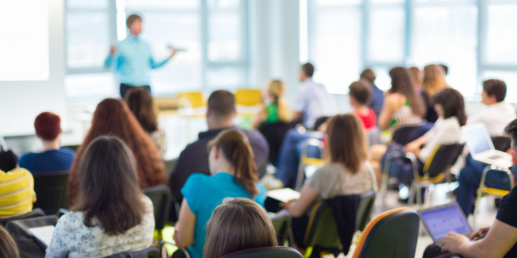 Eine Unterrichtssituation: Im Vordergrund sieht man die Schüler:innen in Reihen an ihren Tischen sitzen, im Hintergrund sieht man den Lehrer, während er etwas erklärt.