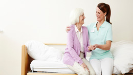 Pflegerin und Pflegebedürftige sitzen auf einem Bett und lächeln sich an.