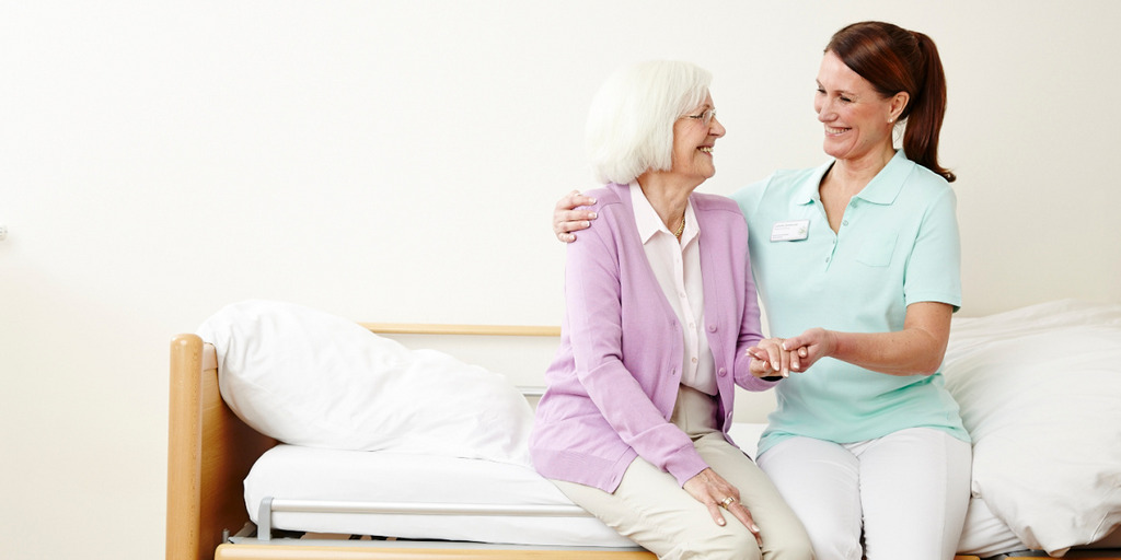 Pflegerin und Pflegebedürftige sitzen auf einem Bett und lächeln sich an.
