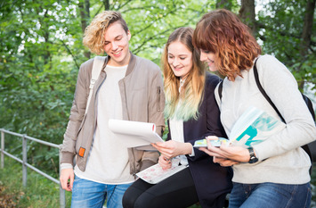 In einem Park stehen drei junge Menschen an ein Geländer gelehnt und blicken gemeinsam auf einen Papierblock, den eine der jungen Frauen in der Hand hält