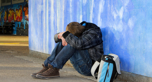 Ein Jugendlicher sitzt auf der Erde, legt seinen Kopf auf die Knie und versteckt sein Gesicht dadurch.