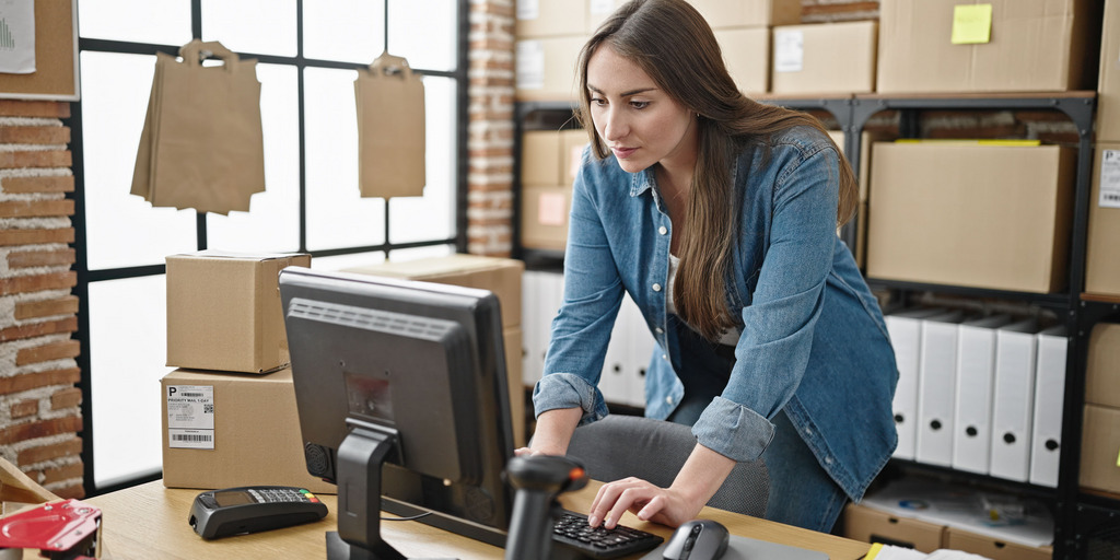 Eine junge Frau beugt sich über einen Schreibtisch, um am PC etwas einzugeben. Im Hintergrund Pappkartons und Aktenordner