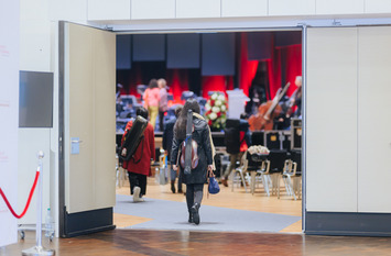 Zwei Musikerinnen betreten, mit ihren Musikinstrumenten auf ihren Rücken, am 10. Oktober 2024 den großen Saal der Stadthalle Gütersloh für die Orchesterprobe zu den NEUEN STIMMEN 2024. Im Saal sieht man Stuhlreihen und im Hintergrund die dekorierte Bühne, auf der drei Personen stehen.