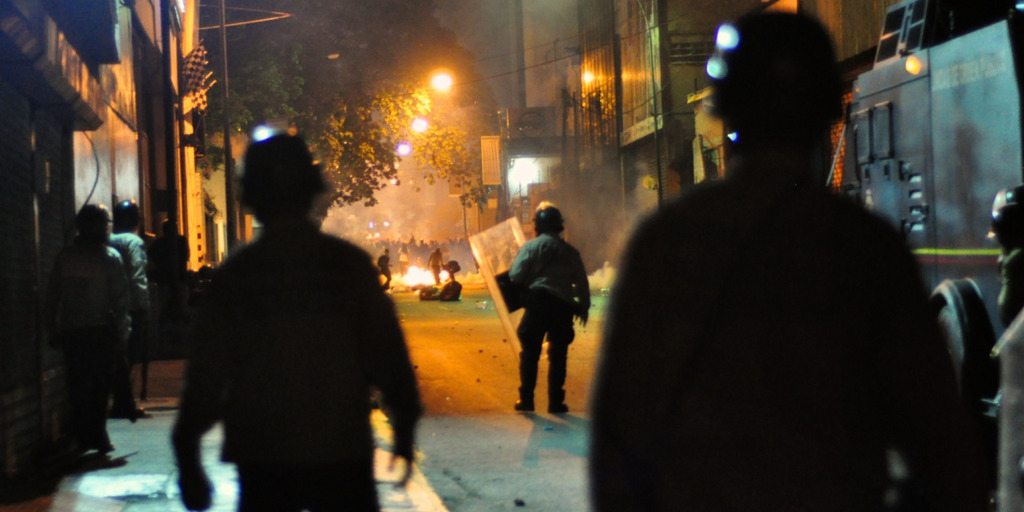On a street of the Venezuelan capital Caracas, soldiers wearing helmets and shields are watching a protesting group of people in the distance. Between them and the protesters there is a fire burning on the street. It is night, a few street lights illuminate the street.