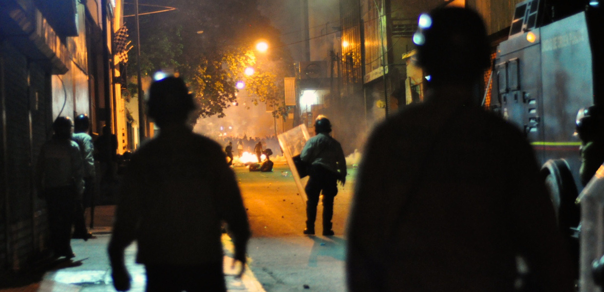 On a street of the Venezuelan capital Caracas, soldiers wearing helmets and shields are watching a protesting group of people in the distance. Between them and the protesters there is a fire burning on the street. It is night, a few street lights illuminate the street.