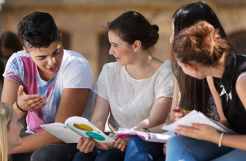 Schülerinnen und Schüler sitzen in einer Gruppe im Freien. Sie halten Bücher und Schreibblöcke in ihren Händen, diskutieren und lachen.