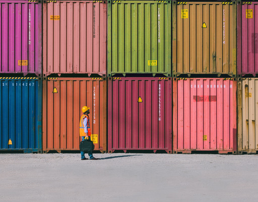 Mehrere bunte Schiffscontainer sind ordentlich übereinandergestapelt, davor geht eine Person in Warnweste und Schutzhelm über eine große, leere Fläche.