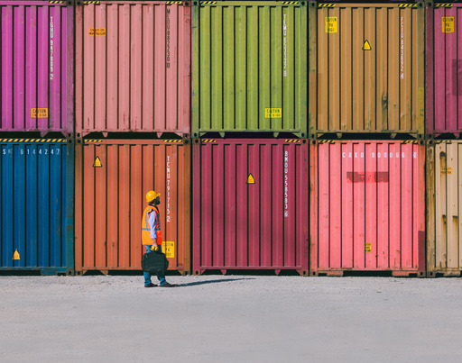 Mehrere bunte Schiffscontainer sind ordentlich übereinandergestapelt, davor geht eine Person in Warnweste und Schutzhelm über eine große, leere Fläche.