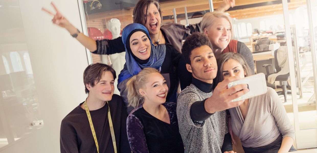 (© martinedoucet/iStockphoto.com) Eine Gruppe von nicht-muslimischen und muslimischen jungen Menschen steht in einem Büro zu einem Selfie zusammen und lacht in die Handykamera.