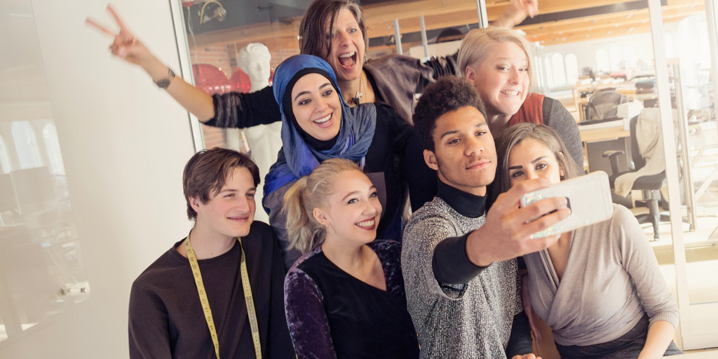 Eine Gruppe von nicht-muslimischen und muslimischen jungen Menschen steht in einem Büro zu einem Selfie zusammen und lacht in die Handykamera.