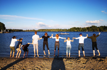 Jugendliche halten sich an der Hand und springen in die Höhe. Sie stehen an einem Strand. Im Hintergrund ist ein See zu sehen.