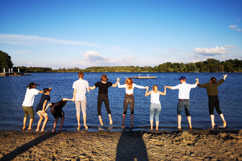 Jugendliche halten sich an der Hand und springen in die Höhe. Sie stehen an einem Strand. Im Hintergrund ist ein See zu sehen.