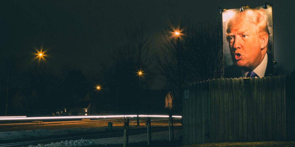 In a backyard at night somewhere in Iowa, a large illuminated sign is towering above the property's fence. The sign is carrying a large photograph of Donald Trump.