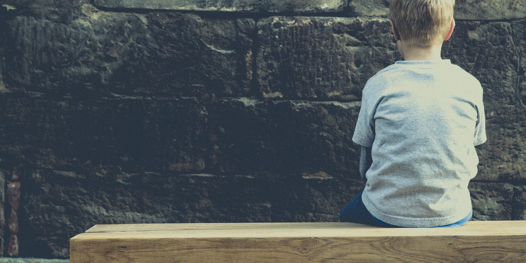 Ein kleiner junge sitzt auf einer Holzbank und starrt auf eine rußschwarze Steinwand eines heruntergekommenen Hauses. Das Kind dreht dem Fotografen den Rücken zu. 