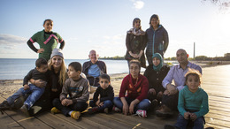 eine Gruppe von geflüchteten Menschen sitzt auf einem Steg am Ufer eines Sees