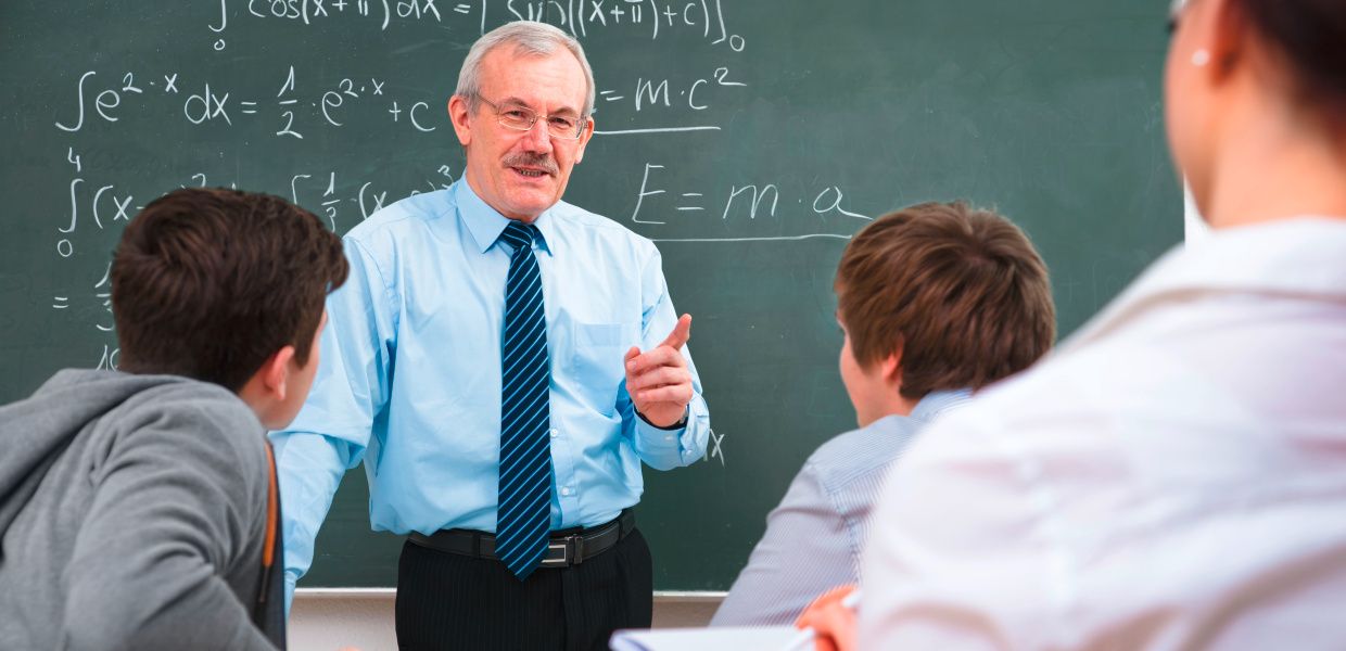 (© Shutterstock / Alexander Raths) Ein Mann steht vor einer Tafel voll von mathematischen Formeln und erklärt drei Personen, die vor ihm sitzen, etwas.