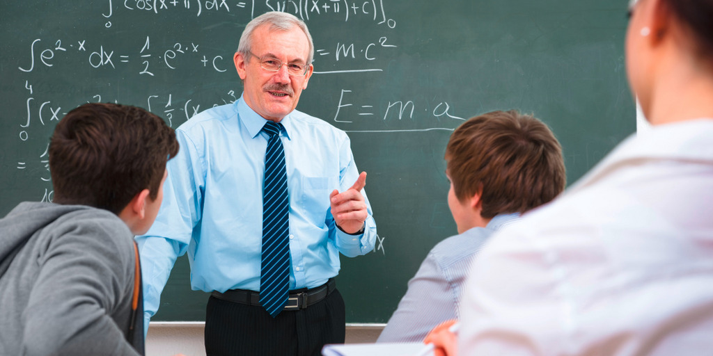 Ein Mann steht vor einer Tafel voll von mathematischen Formeln und erklärt drei Personen, die vor ihm sitzen, etwas.