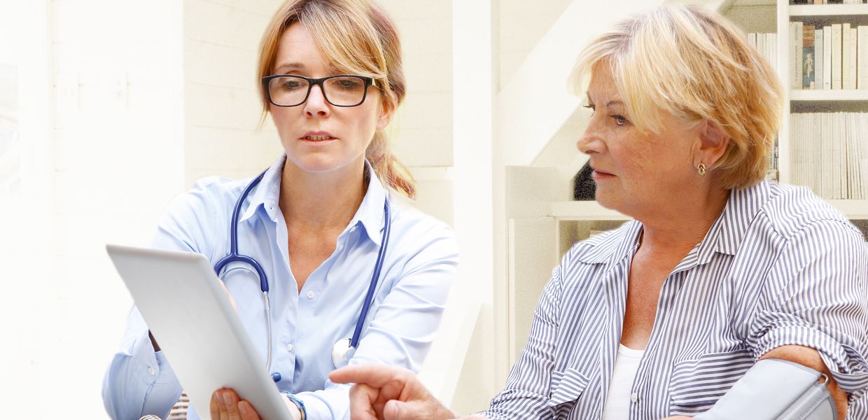 (© Shutterstock/Kinga / Image Source) Ärztin sitzt mit ihrer Patientin am Tisch. Sie schauen gemeinsam auf ein Tablet. Die Patientin hat eine Blutdruckmanschette am Arm.