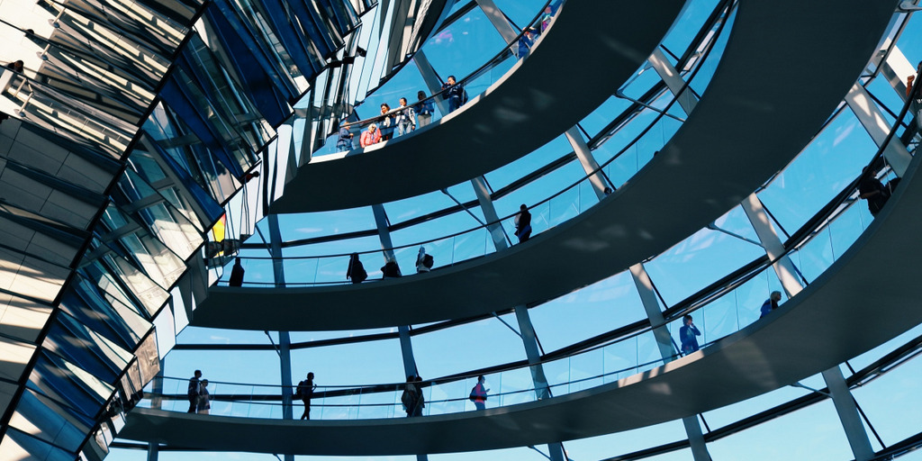 Blick in die Kuppel des Reichstagsgebäudes in Berlin. Vor einem blauen Himmel gehen Besucher:innen die spiralförmig gewundene Rampe zur Spitze der Kuppel hinauf.